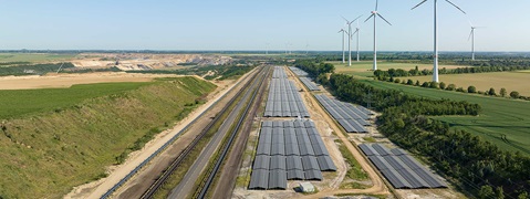 Luftaufnahme einer Landschaft mit Windkraftanlagen und Solarmodulen entlang der Eisenbahnlinie.