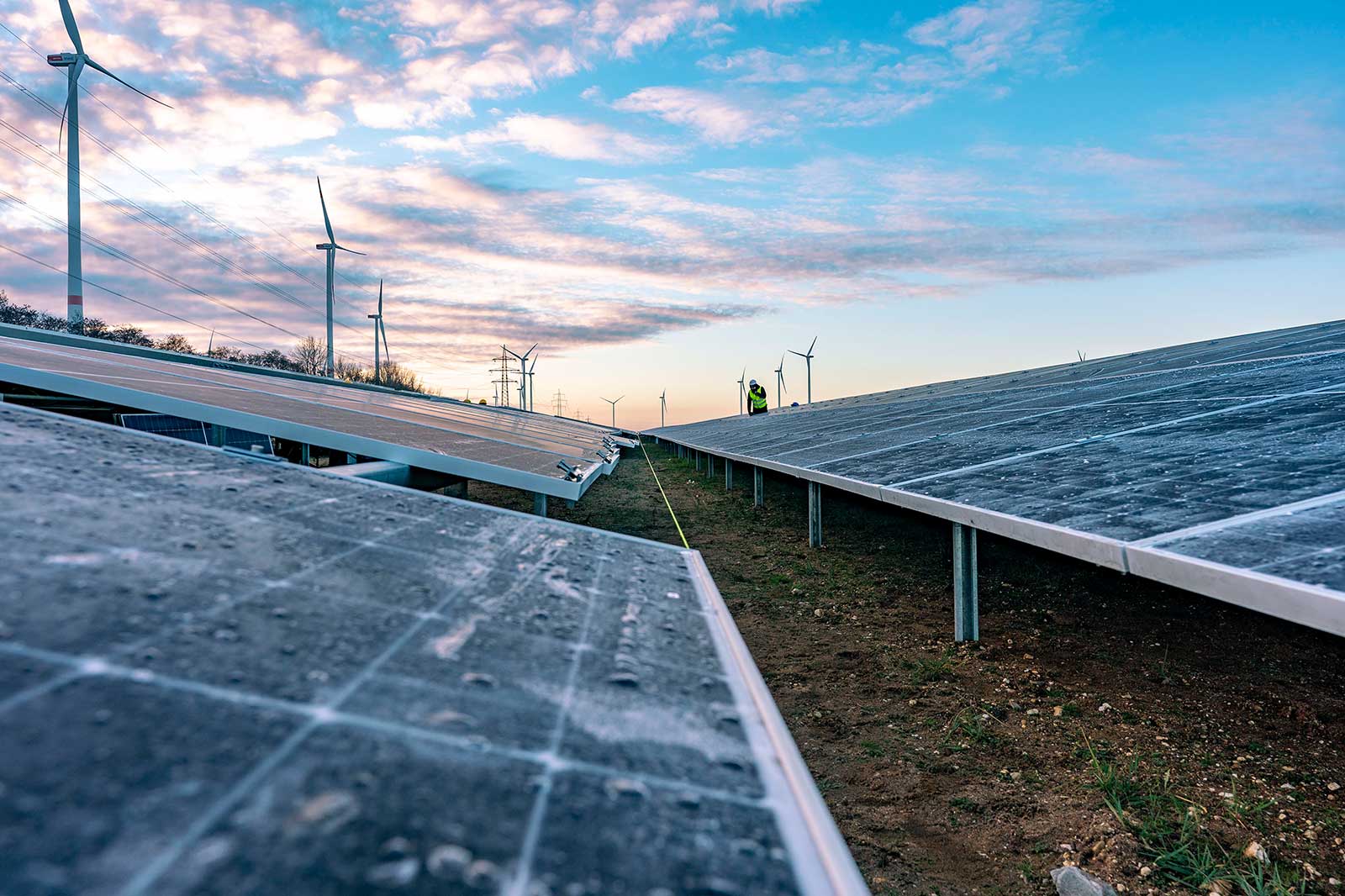 Ein Solarfeld mit Solarpanels im Vordergrund und Windturbinen im Hintergrund unter einem bunten Himmel bei Sonnenuntergang.