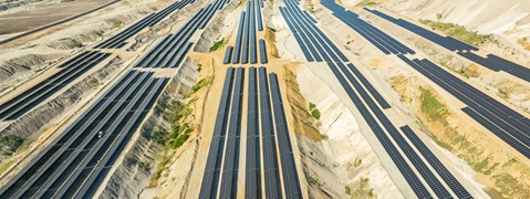Ein Luftbild einer Solaranlage mit zahlreichen Solarpanels, die in schmalen Reihen in einem sandigen Gelände angeordnet sind.