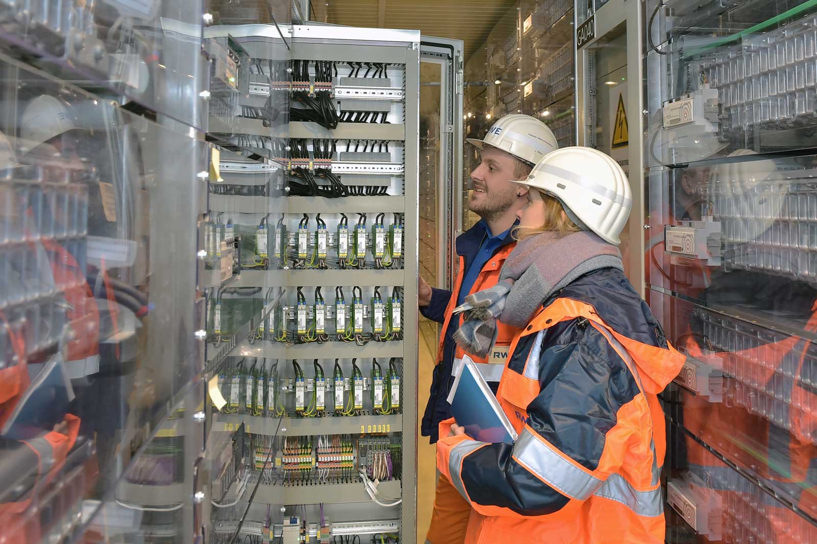 Two workers in safety helmets inspect electrical equipment in a facility, surrounded by cabinets filled with circuits and cables.
