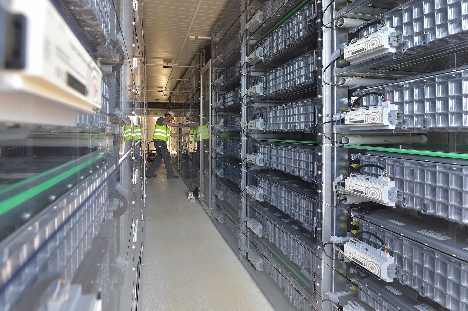 An interior space filled with shelves of batteries. Two workers in safety vests are attending to the equipment.