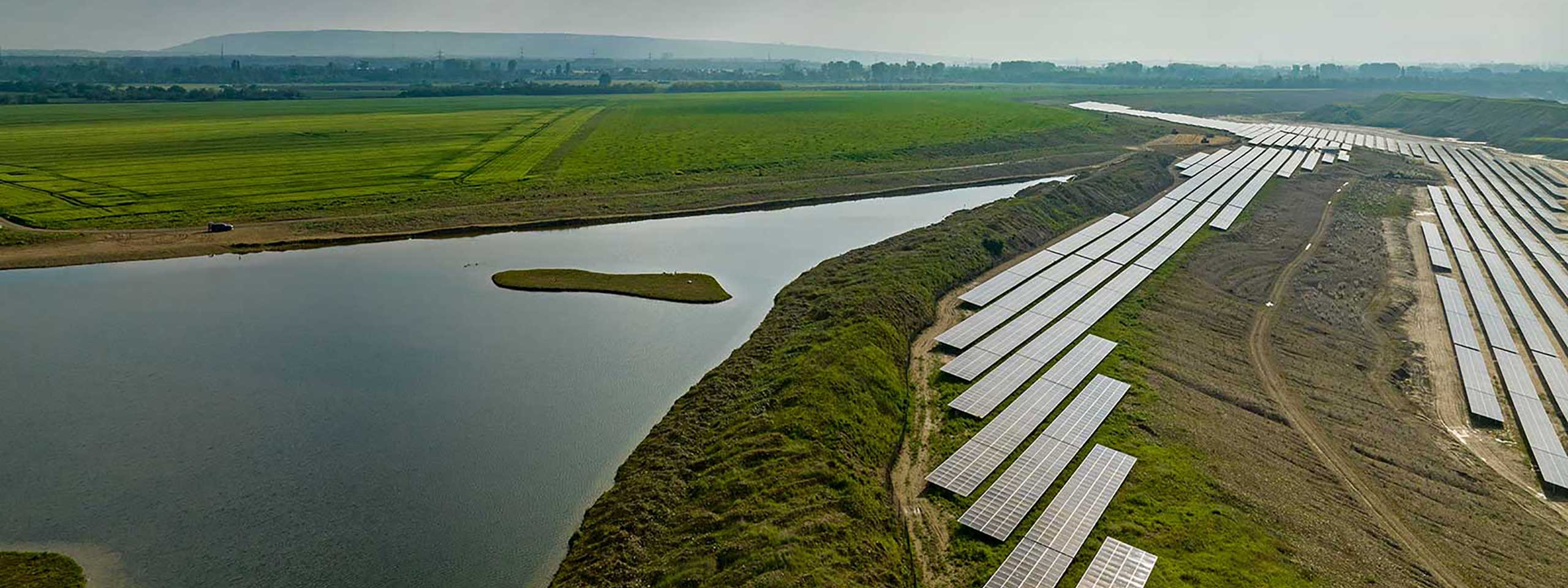 Eine große Solaranlage neben einem ruhigen Gewässer, umgeben von grünen Feldern und sanften Hügeln im Hintergrund.