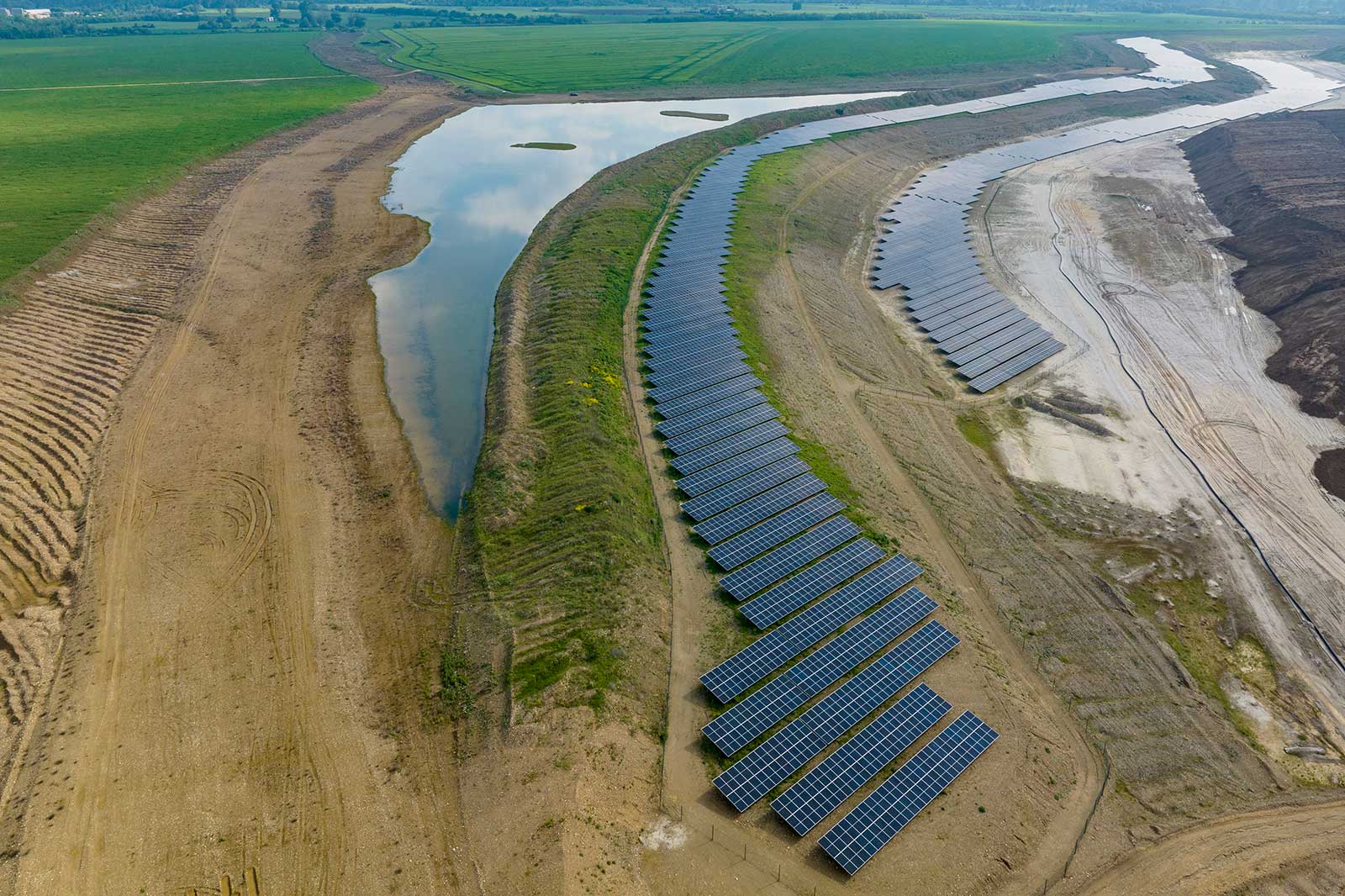 Luftaufnahme von Solarpanels, die entlang eines gebogenen Uferbereichs in einer grünen Landschaft angeordnet sind.