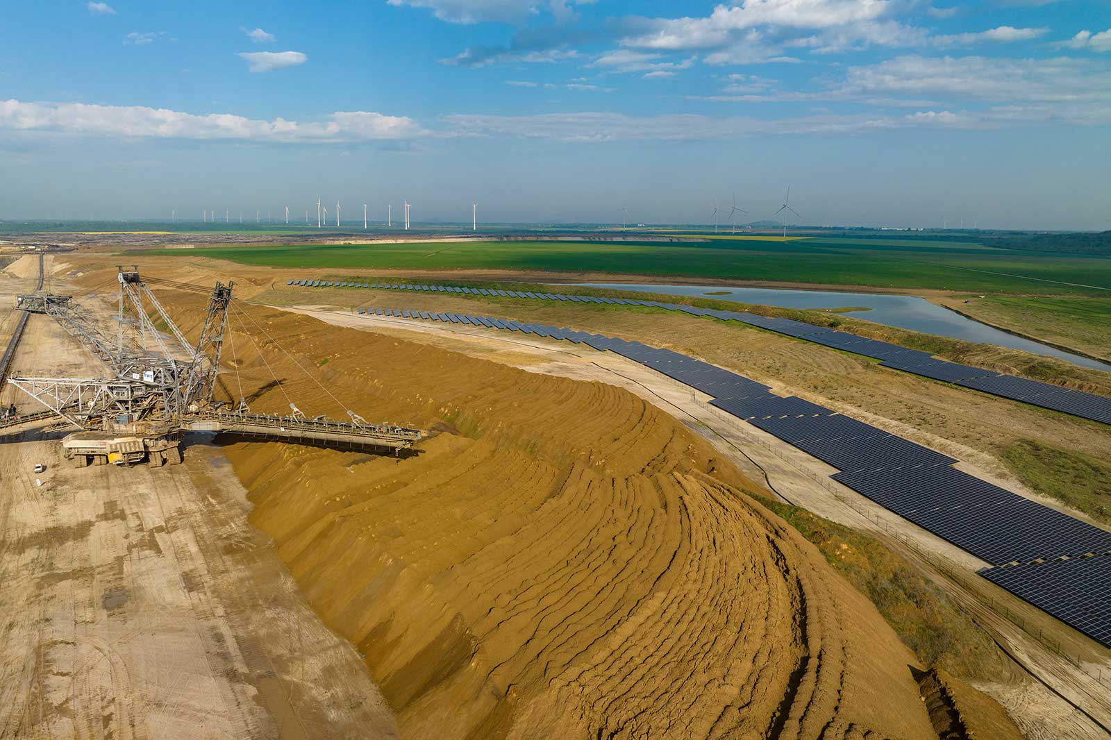 Eine Landschaft mit Solarpanelen und Windkraftanlagen. Ein großer Bagger arbeitet in einer aufgeschütteten Grube.