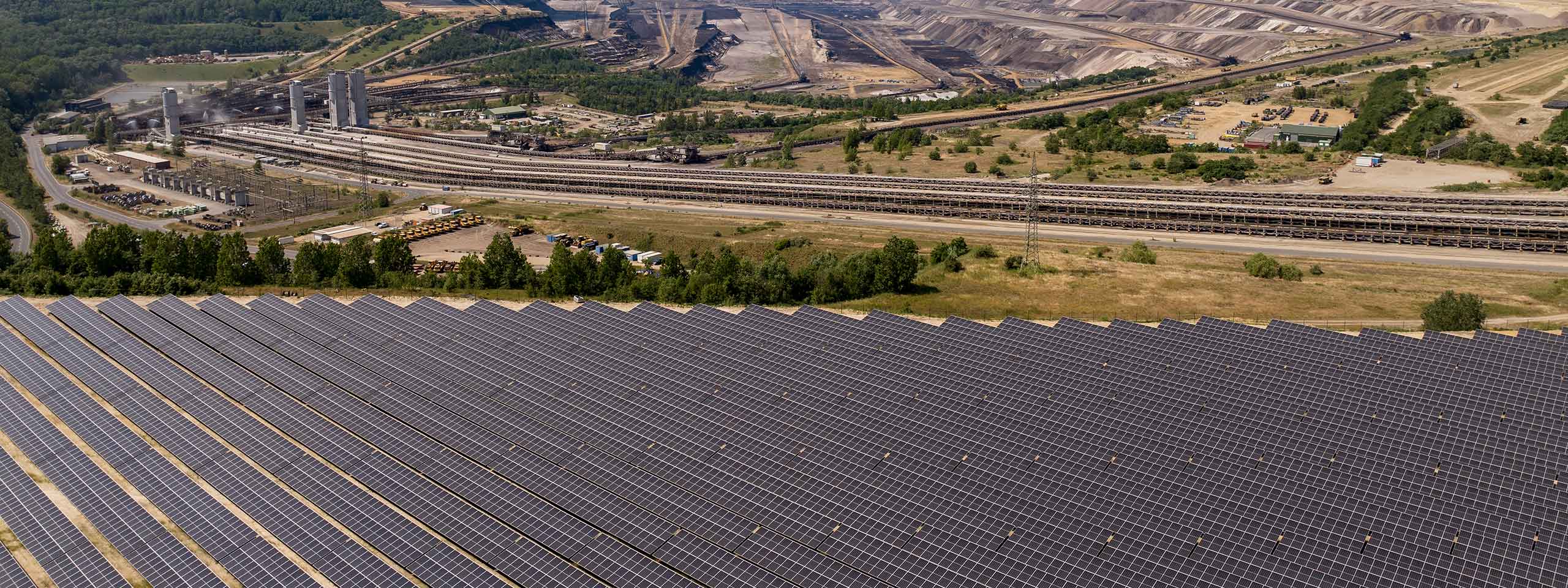 A large solar panel field in the foreground with an industrial facility in the background, surrounded by trees and hills.