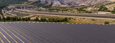 Ein großes Solarpanelfeld in der Vordergrund mit einer Industrieanlage im Hintergrund, umgeben von Bäumen und Hügeln.