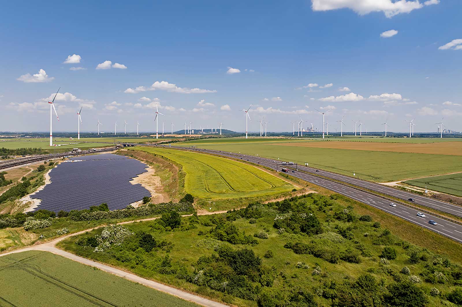 Landschaft mit Windkraftanlagen und Solarpanelen, grüne Felder und eine Straße unter blauem Himmel.