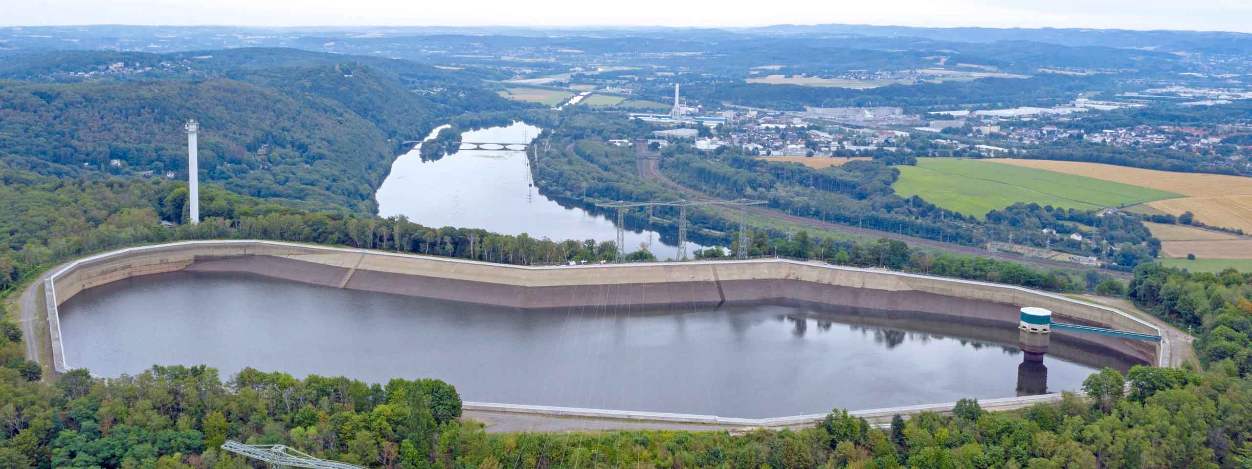 Luftaufnahme eines großen Stausees, umgeben von Wäldern, mit einem Reservoir und einem hohen Turm im Hintergrund.