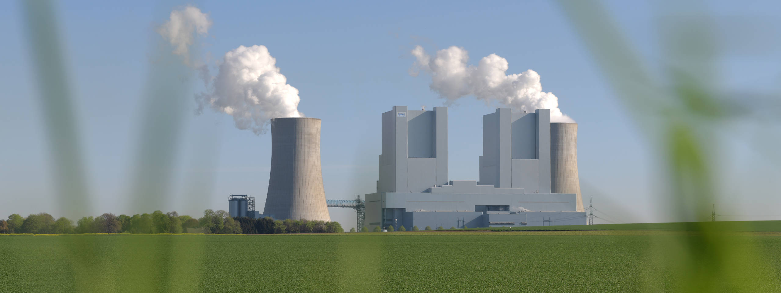 Ein Kraftwerk mit Kühltürmen und rauchenden Schornsteinen, umgeben von grünem Feld und blauen Himmel.