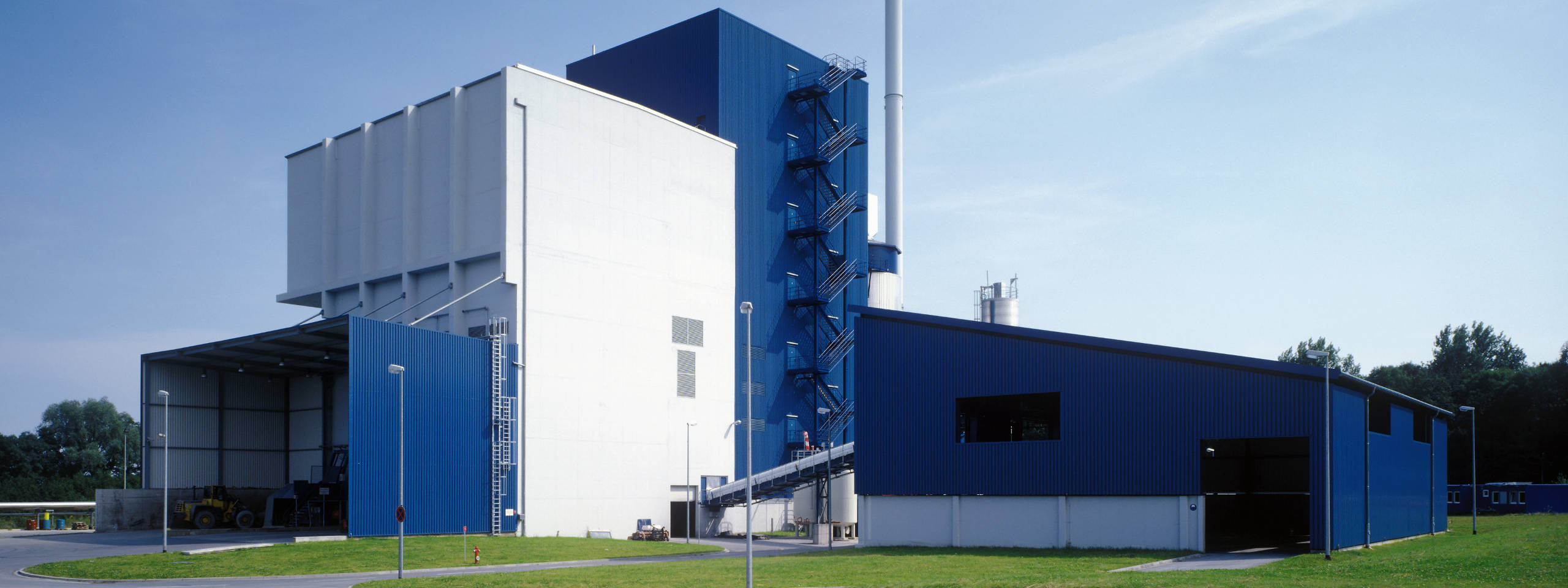 Ein modernes Industriegebäude mit blauer Fassade, großen Fenstern und einem hohen Schornstein vor einem klaren Himmel.
