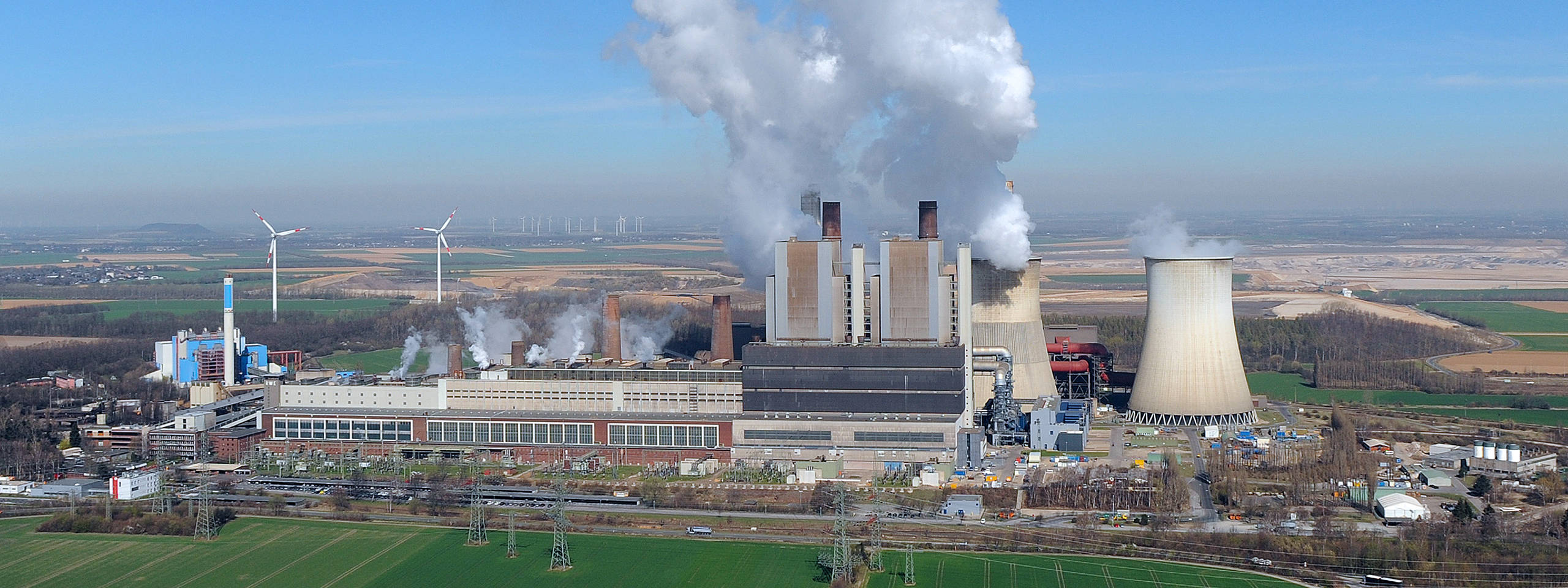Ein Industriekomplex mit rauchenden Schornsteinen, Kühltürmen und Windkraftanlagen vor einer ländlichen Kulisse.