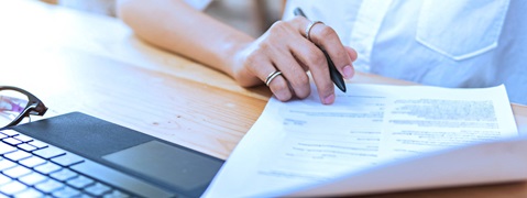 Eine Person hält einen Stift über ein Dokument, während sie an einem Holztisch sitzt, mit einem Laptop und einer Brille in der Nähe.