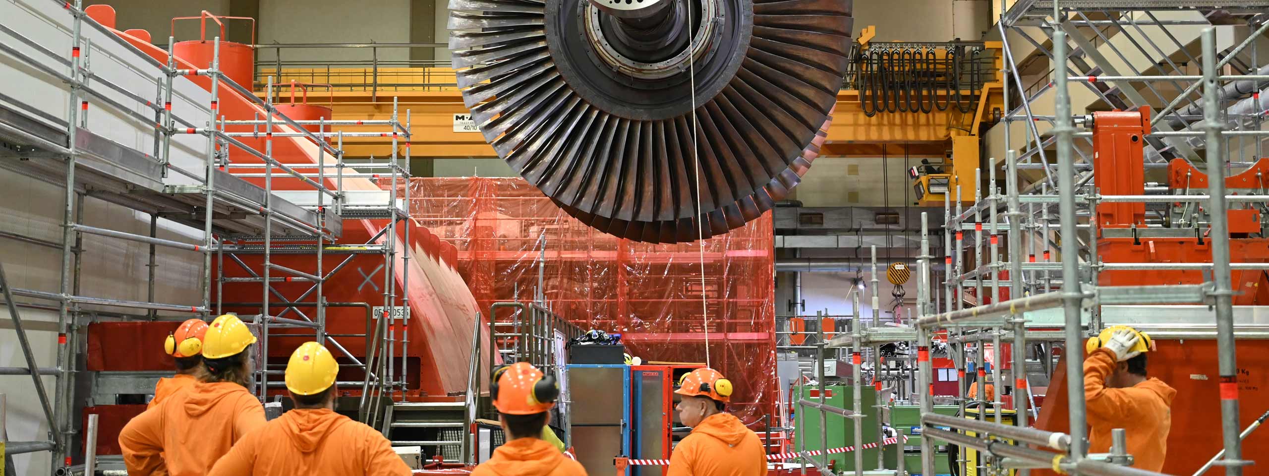 Ingenieure arbeiten in einer großen Fabrikhalle, während eine Turbine über ihnen hängt, umgeben von Gerüsten und Sicherheitsmaßnahmen.