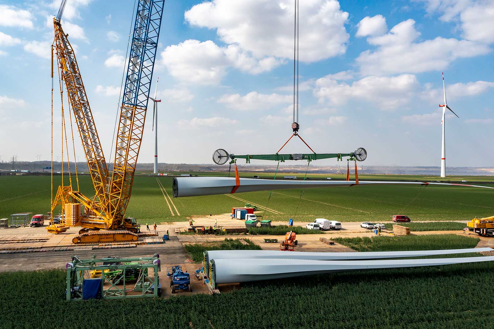 Ein grosser Kran hebt Windturbinenblätter auf einem grünen Feld, mit weiteren Windturbinen im Hintergrund und Bauarbeitern.