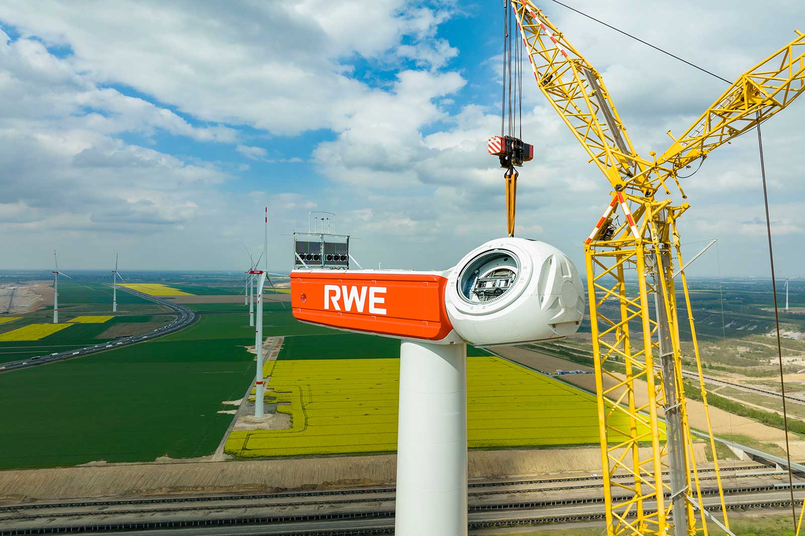 Eine Windkraftanlage mit einem orangefarbenen RWE-Logo, Kran und grünen Feldern im Hintergrund unter einem bewölkten Himmel.