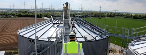 Ein Arbeiter steht auf einem großen Behälter, umgeben von einer landschaftlichen Aussicht und Hochspannungsleitungen.