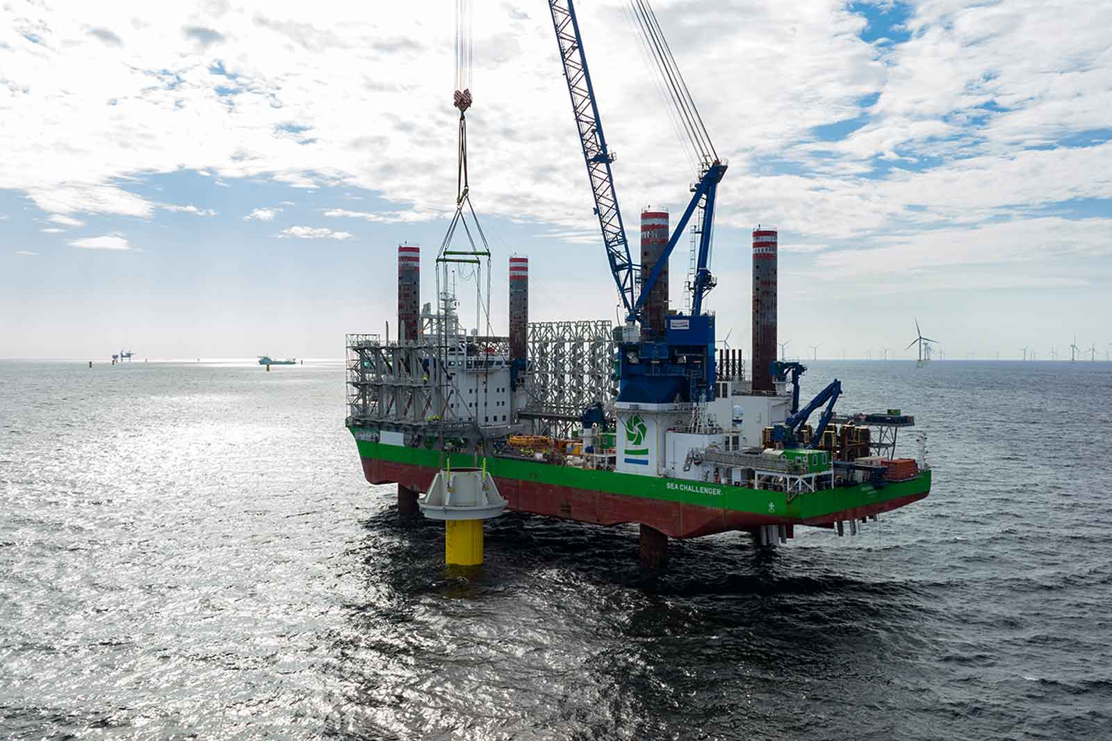 Ein Offshore-Schiff mit einem großen Kran hebt eine gelbe Struktur aus dem Wasser, umgeben von Windkraftanlagen im Hintergrund.