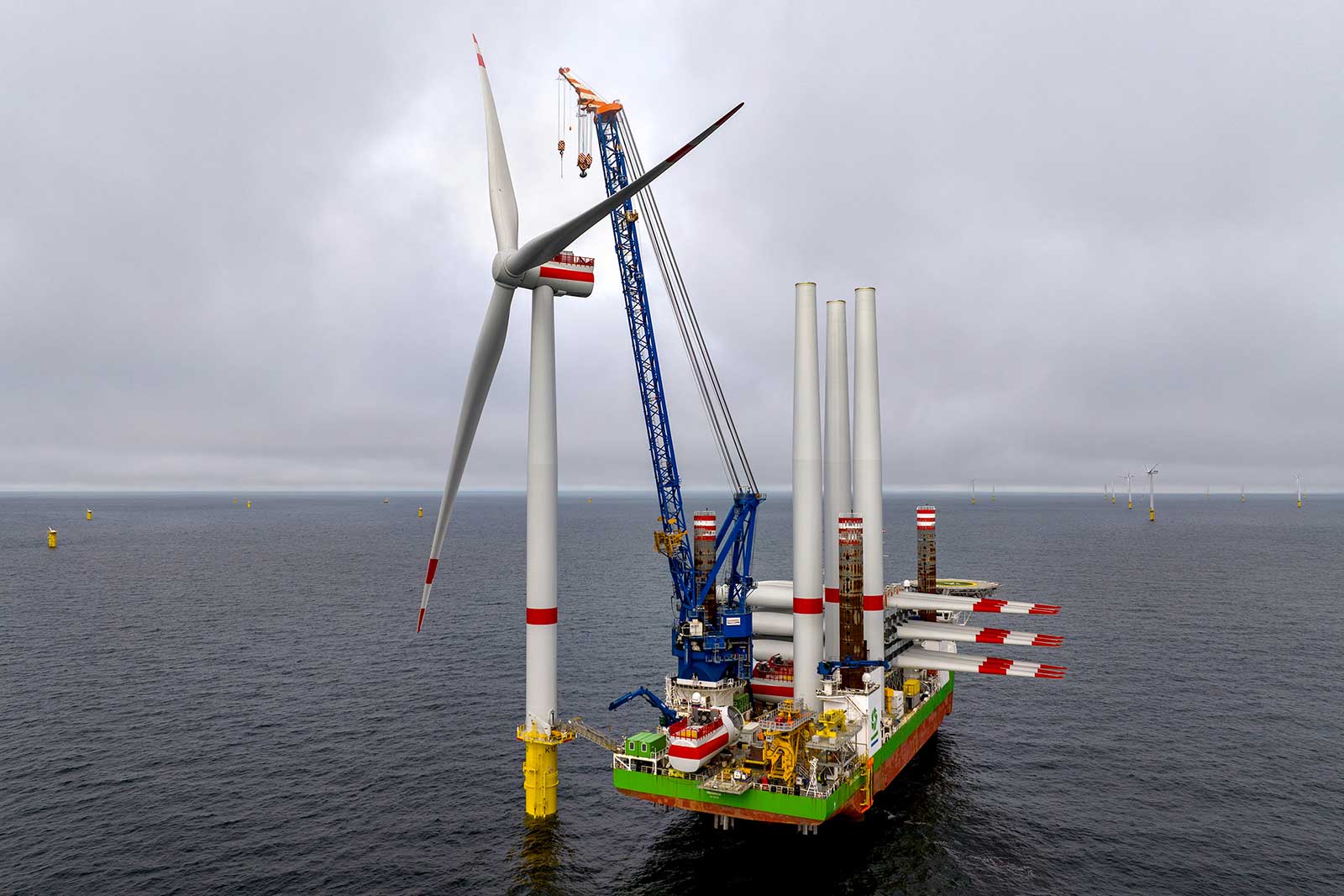 Ein großes Offshore-Windkraftanlagenmontageschiff mit einem Kran, in der Nähe von Windturbinen im Meer unter einem bewölkten Himmel.