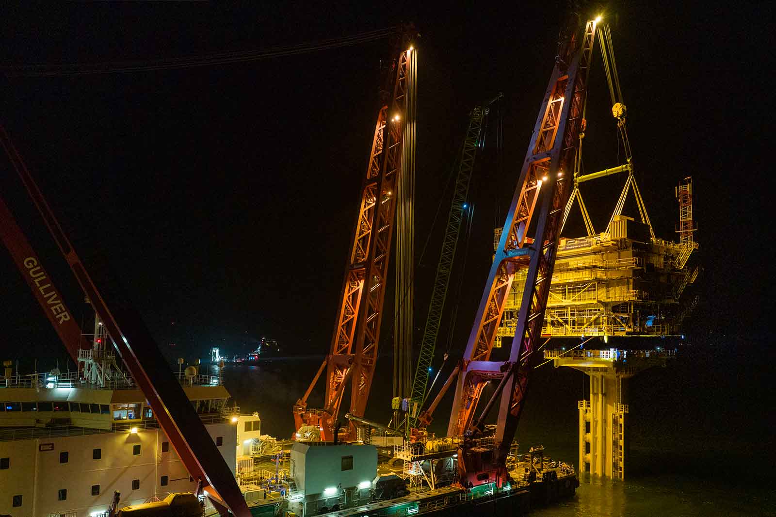 Ein Offshore-Schiff hebt in der Nacht eine Plattform mit orangenen Kränen an. Das Wasser reflektiert die Lichter.