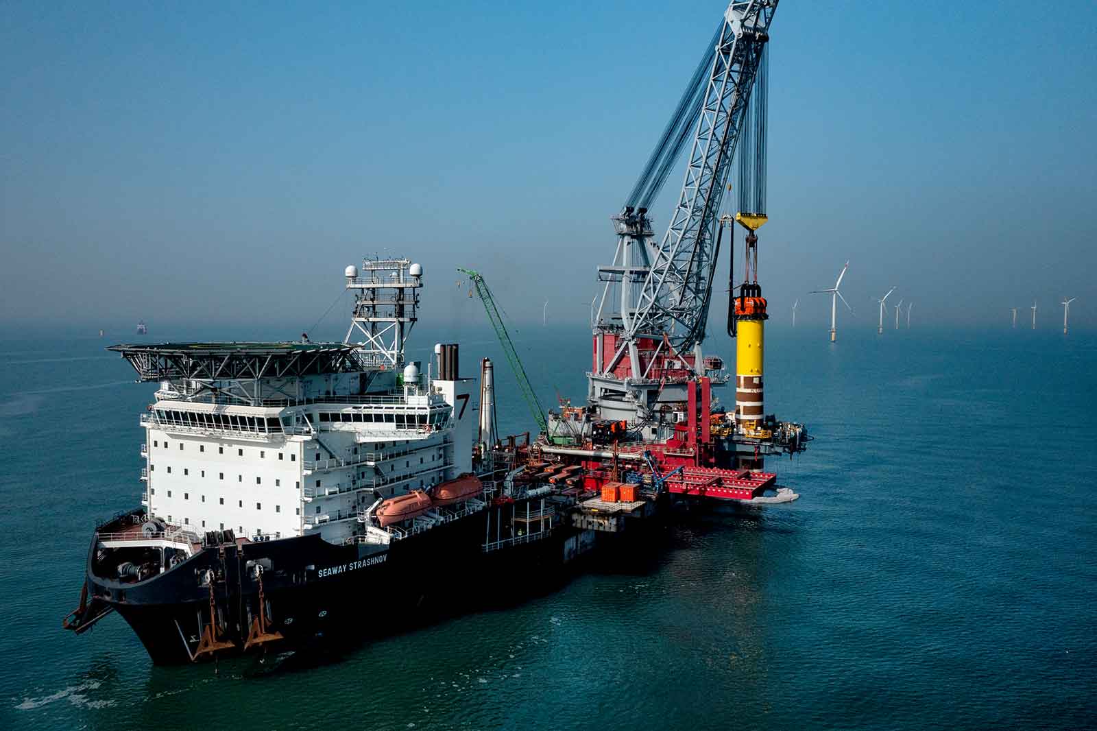 Ein großes Schiff mit einem Kran, das im Meer arbeitet, mit Windkraftanlagen im Hintergrund sichtbar.