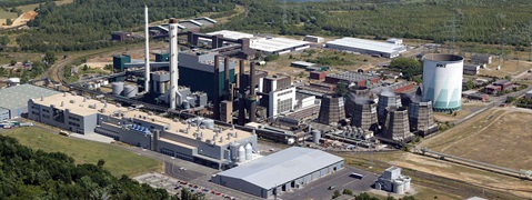 Luftaufnahme eines großen Industriekomplexes mit Schornsteinen, Lagertanks und Kühltürmen, umgeben von Grünflächen und Straßen.