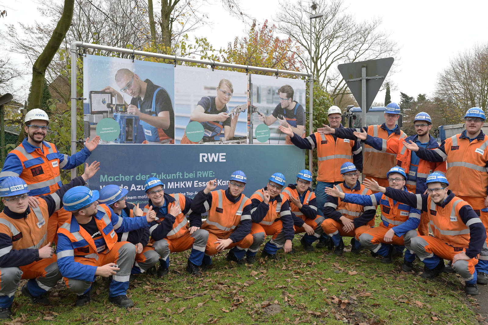 Gruppe von Arbeitern in orange-blauen Uniformen, die im Freien zusammen posieren, mit einem Werbebanner im Hintergrund.