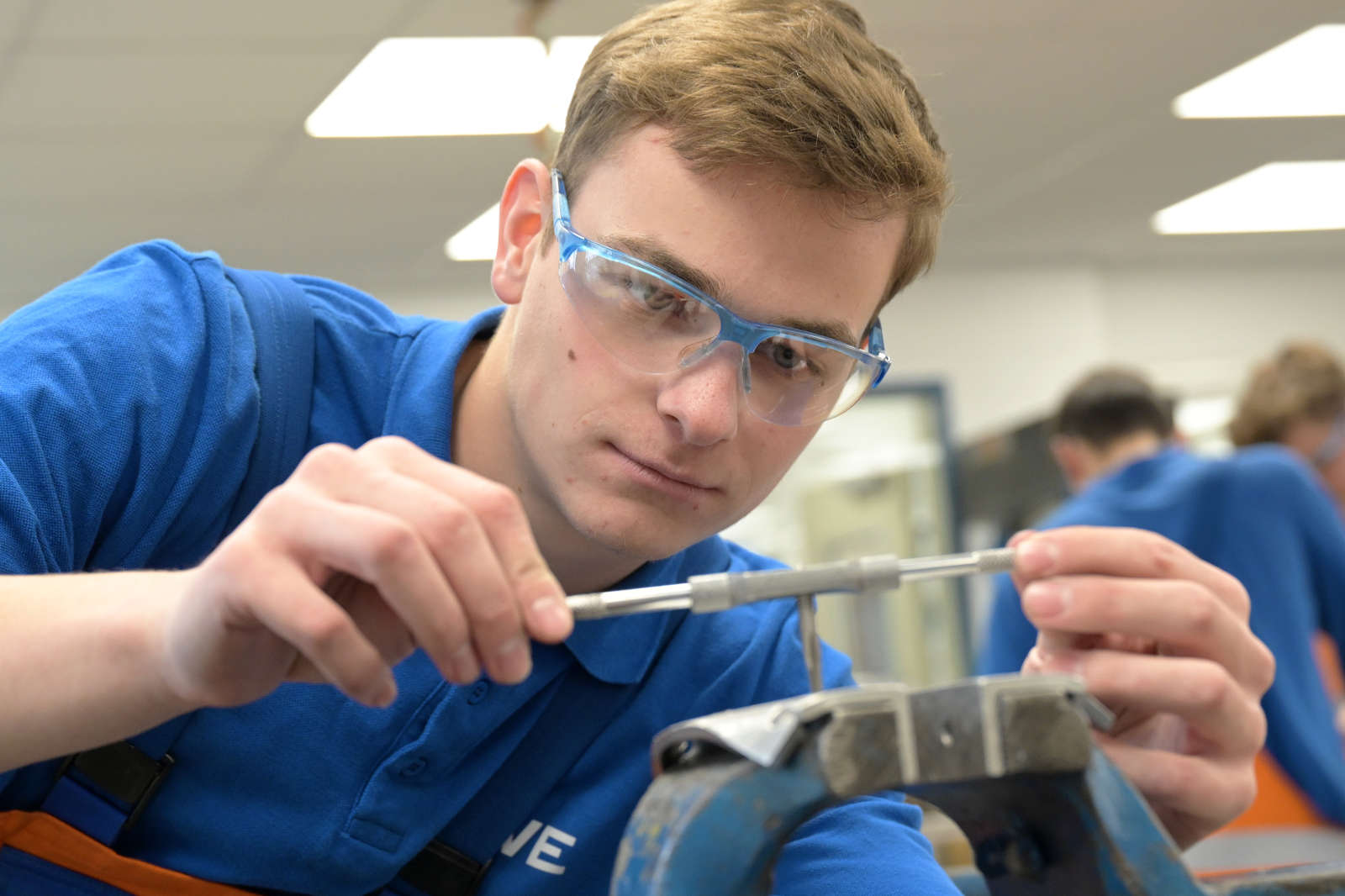 Ein Schüler in einem blauen Hemd benutzt ein Werkzeug, während er in einer Werkstatt arbeitet, konzentriert auf seine Aufgabe mit sichtbaren Werkzeugen.