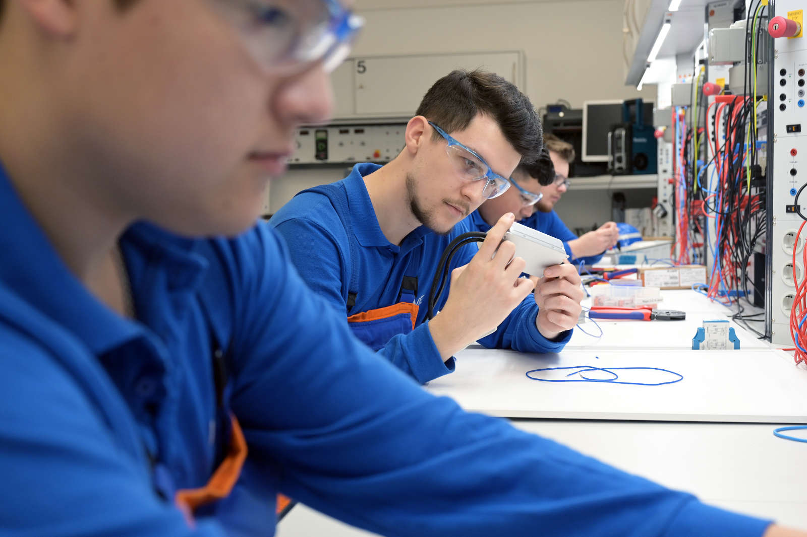 Drei Personen in blauen Uniformen arbeiten an einem Tisch, der mit Werkzeugen und Geräten in einer Werkstatt gefüllt ist.