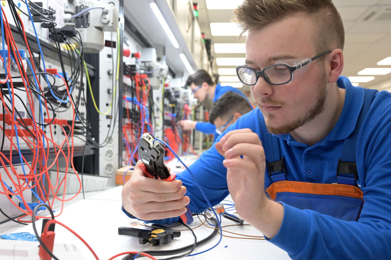 Eine Person, die Werkzeuge und Drähte in einem Arbeitsbereich mit elektrischen Geräten und bunten Kabeln hantiert.