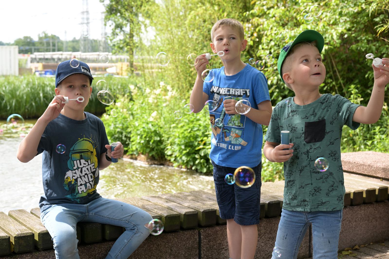 Drei Kinder, die Blasen in einem Garten pusten, umgeben von Grünpflanzen und einem Wasserbecken.
