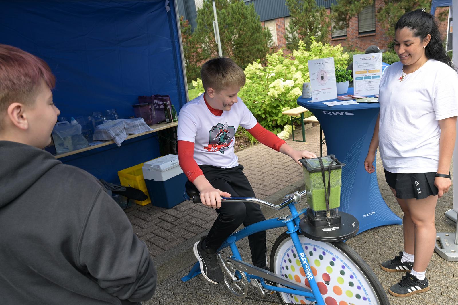 Ein Kind fährt ein stationäres Fahrrad, das mit einem Mixer verbunden ist, um einen grünen Smoothie herzustellen, während andere zuschauen.
