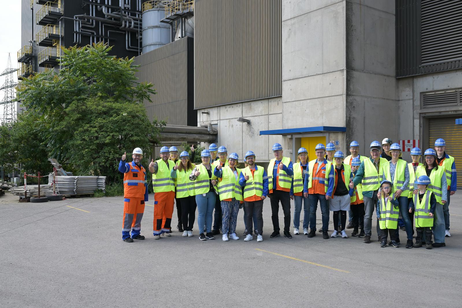 Eine Gruppe von Arbeitern in Sicherheitskleidung steht draußen vor einem Gebäude und lächelt, während sie Daumen hoch zeigen.