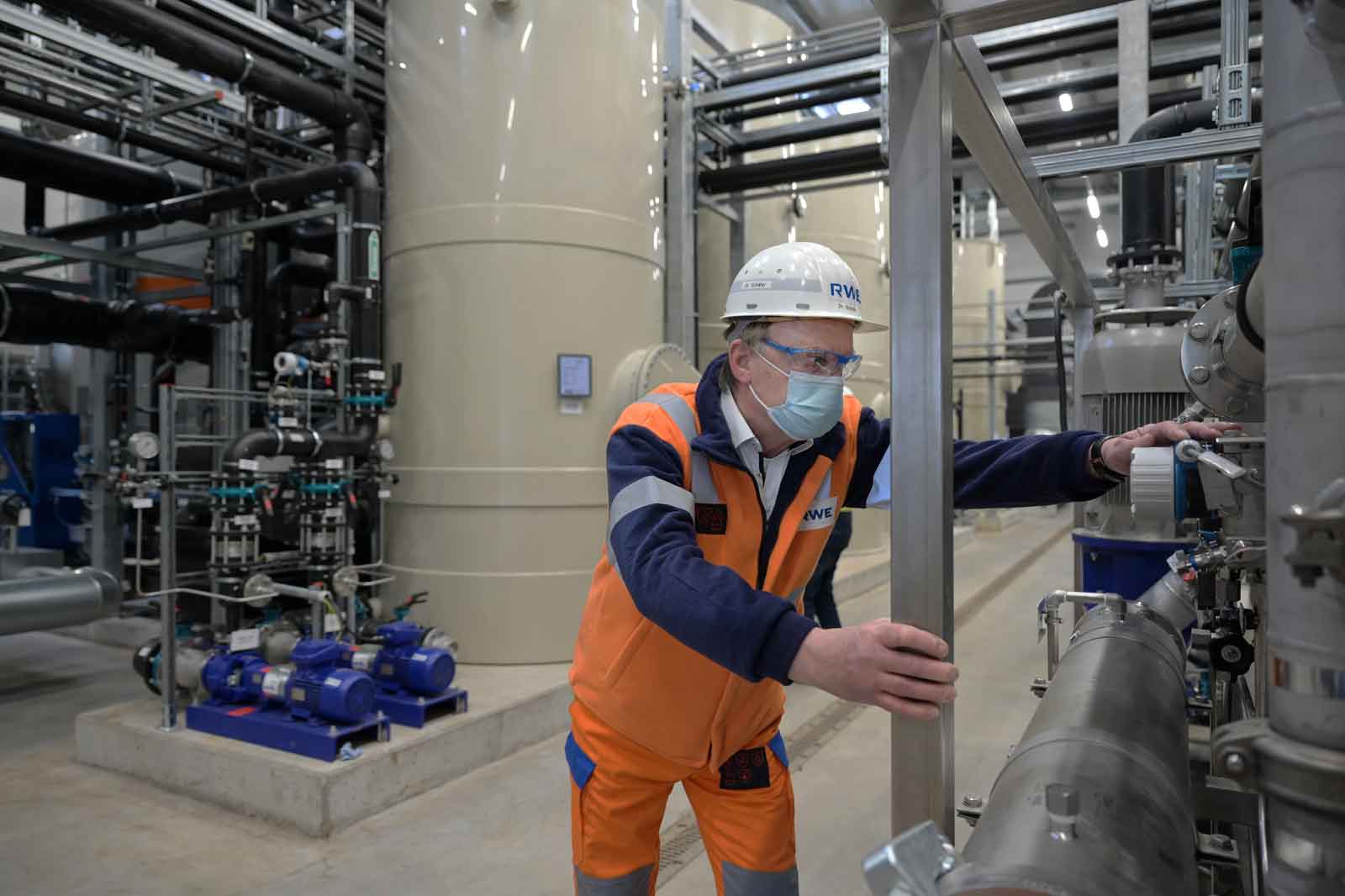 Ein Arbeiter in einer orange-blauen Uniform bedient Maschinen in einer großen Industrieanlage mit Rohren und Pumpen im Hintergrund.