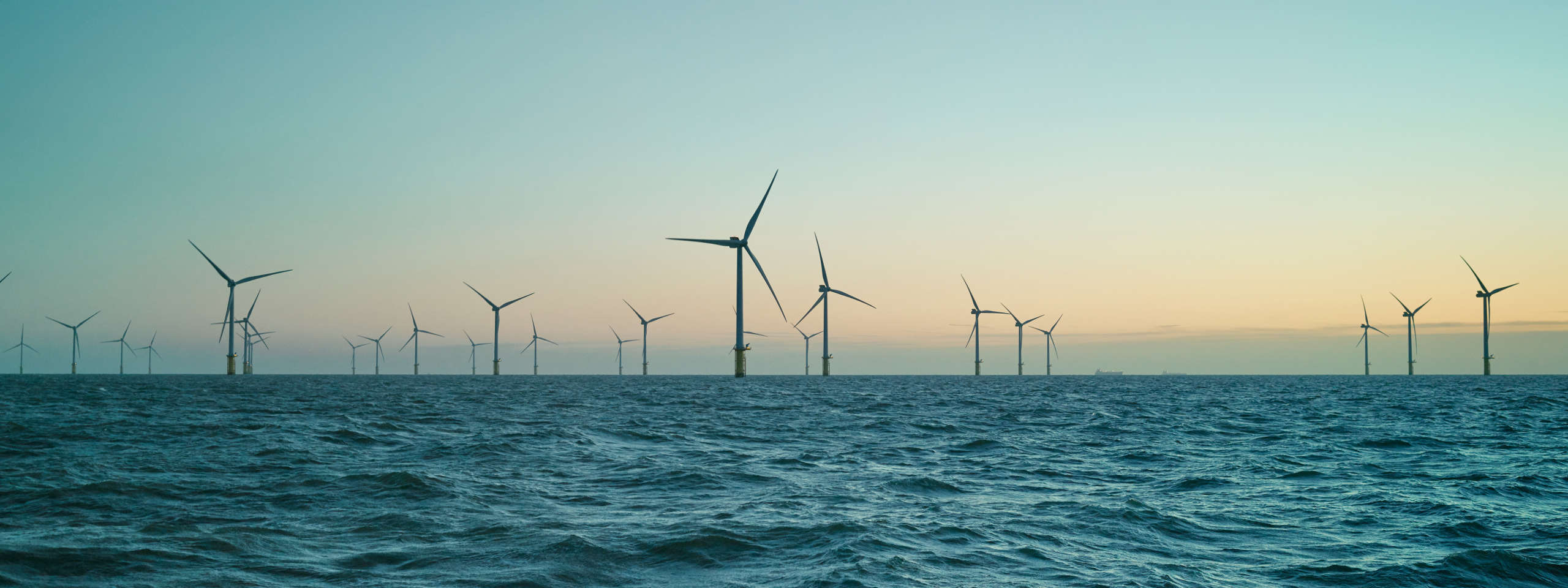 Eine Reihe von Windkraftanlagen steht im Wasser bei Sonnenuntergang, der Himmel hat einen sanften Farbverlauf.