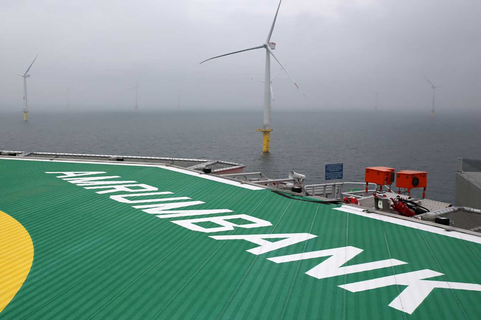 Ein Hubschrauberlandeplatz mit dem Schriftzug AMRIMBANK, im Hintergrund Windturbinen auf dem Wasser in nebliger Umgebung.