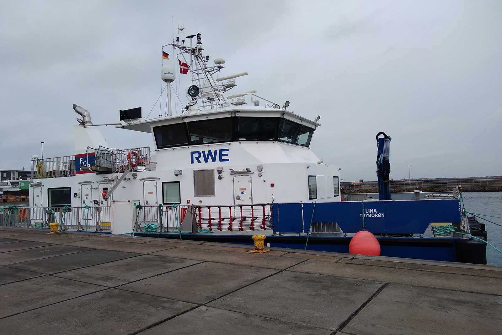 Ein modernes Schiff mit der Aufschrift RWE liegt an einem Hafen, umgeben von grauem Himmel und Hafenanlagen.