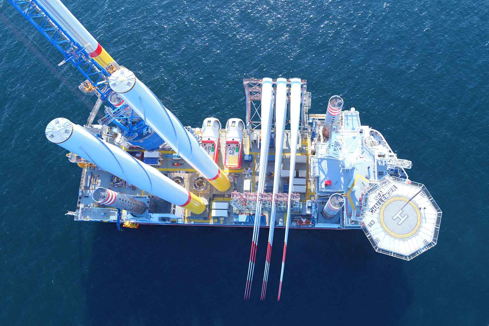 Eine Luftaufnahme eines großen Offshore-Schiffs mit Windturbinenblättern und einem Kran auf dem Deck, umgeben von tiefblauem Wasser.