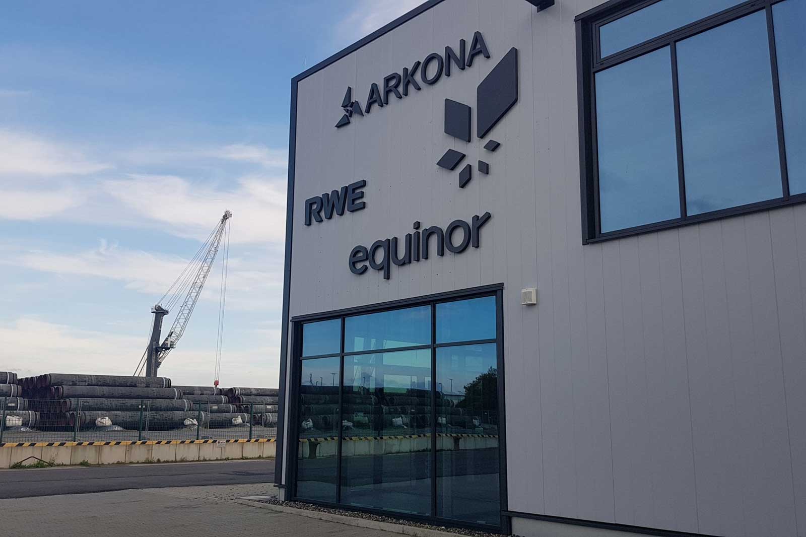 Die Gebäudeaußenansicht mit den Logos von Arkona, RWE und Equinor. Im Hintergrund sind gestapelte Rohre und ein Kran zu sehen.