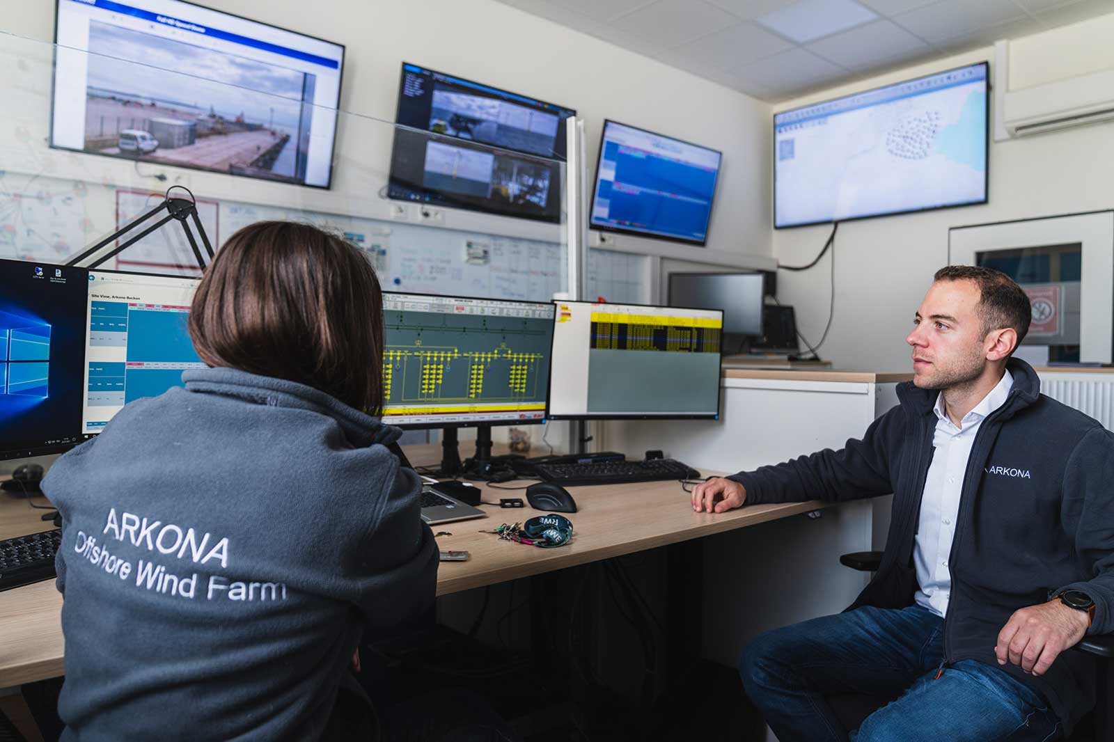 Zwei Personen im Kontrollraum einer Offshore-Windfarm, umgeben von Monitoren und technischen Geräten.