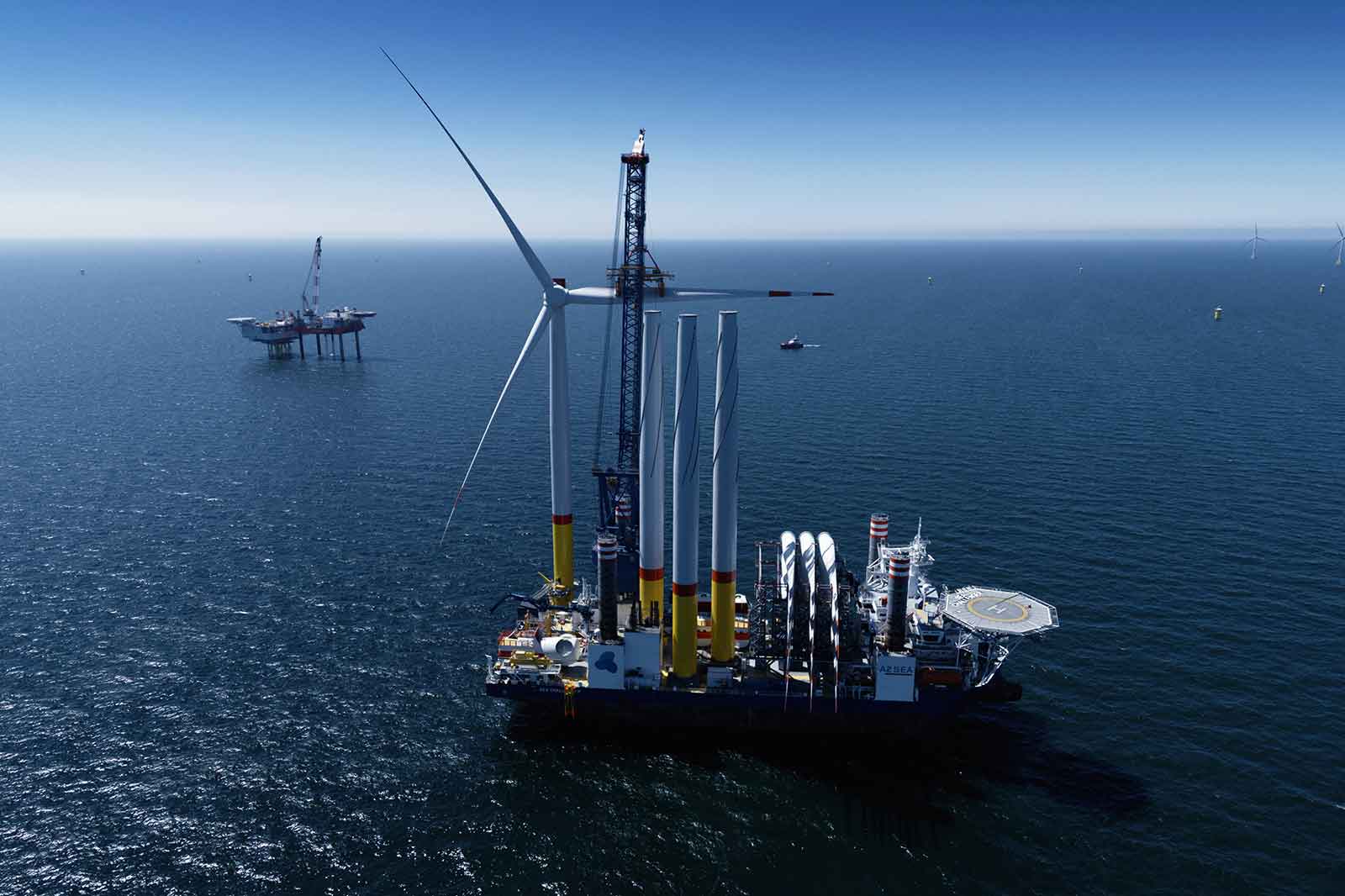 Ein großes Offshore-Windkraftinstallationsschiff mit mehreren Turbinen und einer Bohrplattform im Hintergrund.