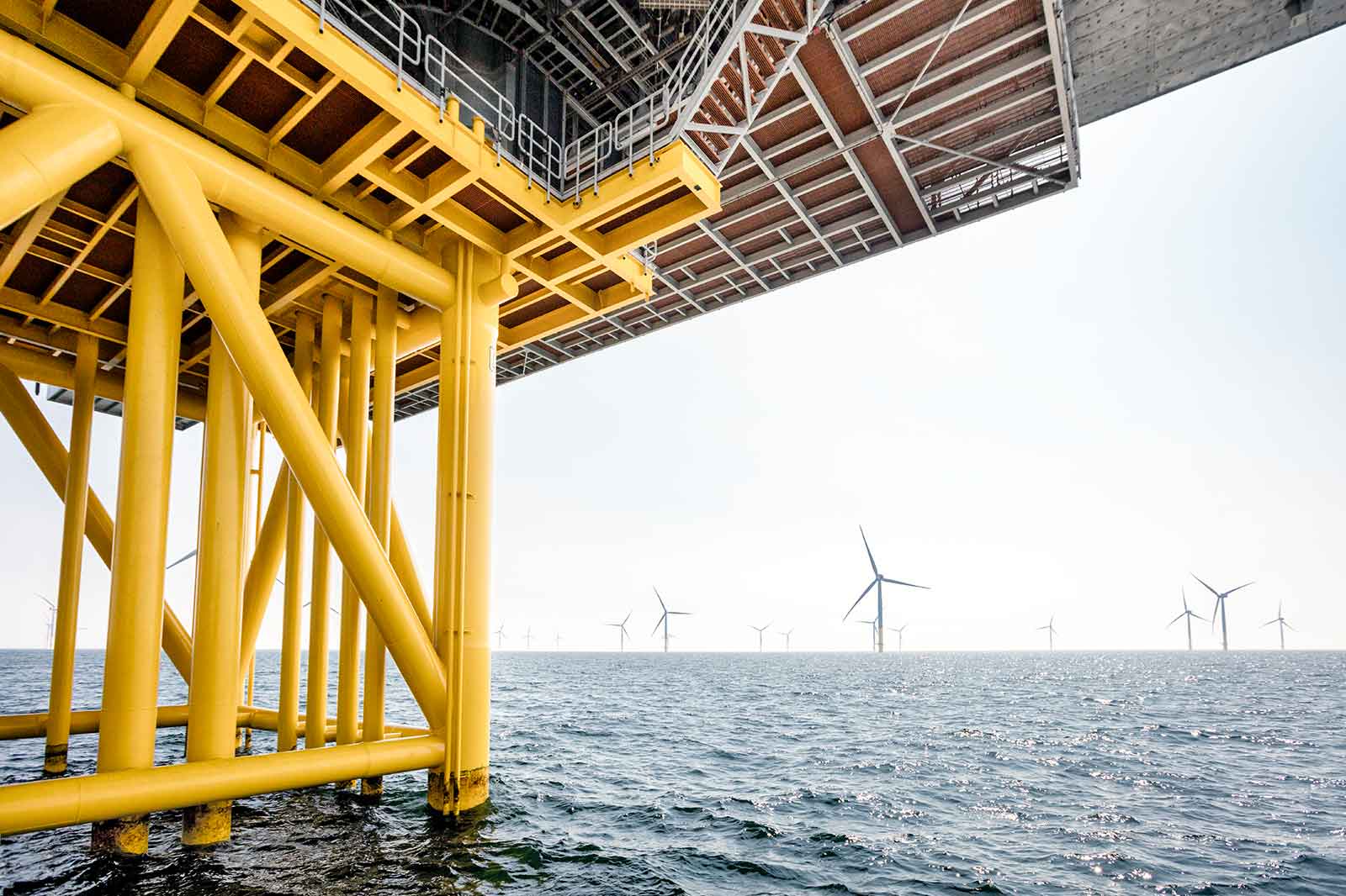 Ein Blick auf die gelbe Unterkonstruktion einer Offshore-Anlage, mit Windturbinen im Hintergrund über dem Wasser.