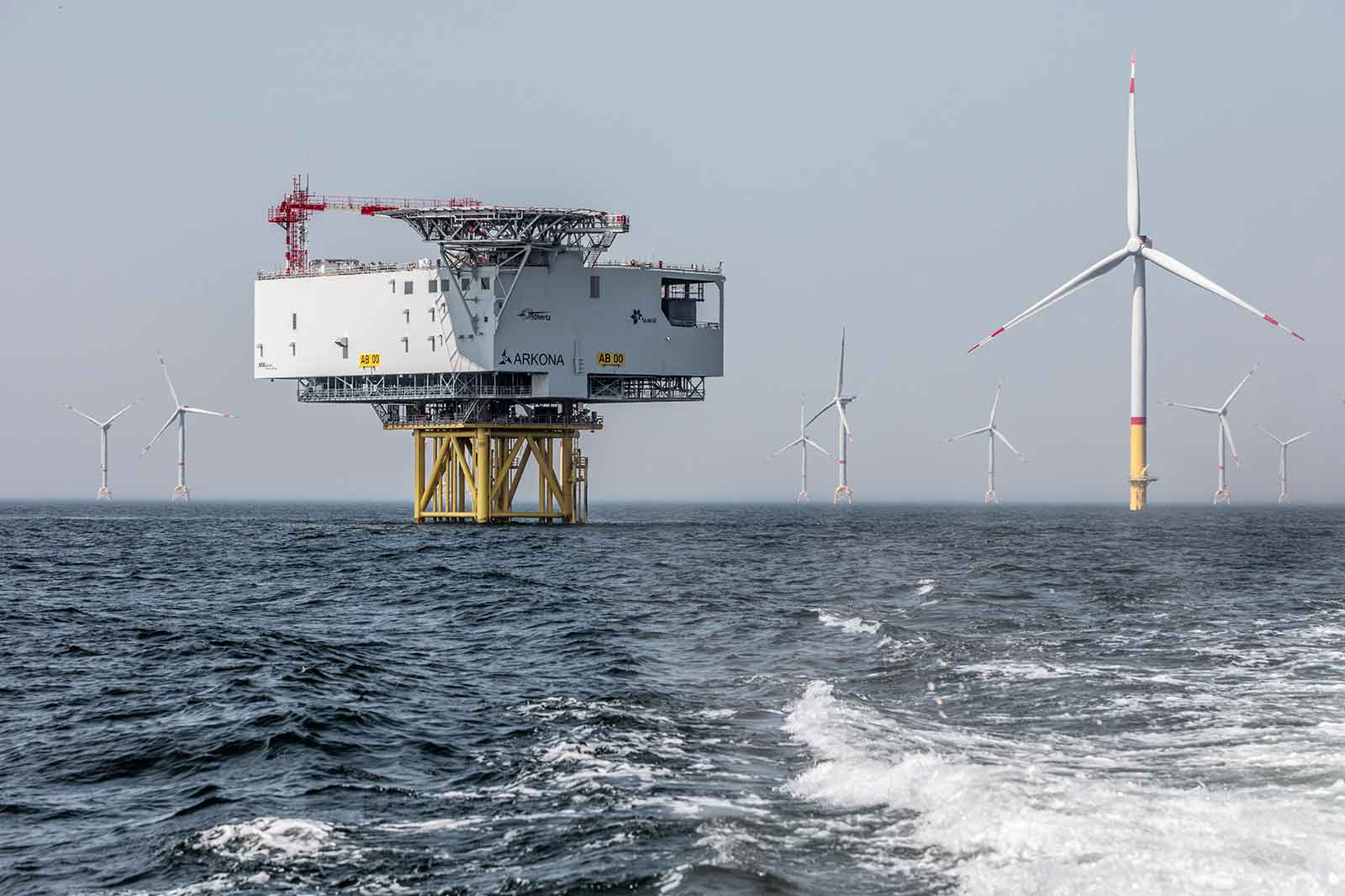 Ein Offshore-Windpark mit einem großen Umspannwerk und mehreren Windturbinen im Hintergrund, die in ruhigem Wasser stehen.