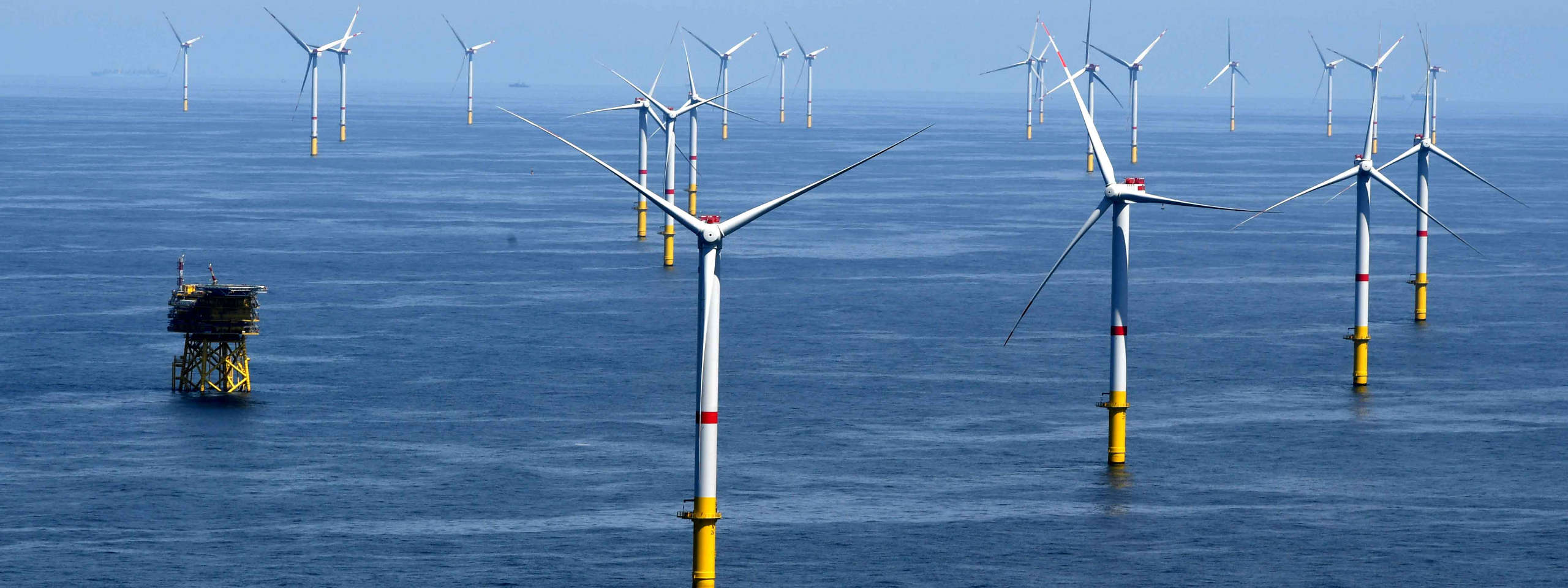Eine Reihe von Windkraftanlagen auf dem Meer vor einem blauen Himmel. Im Vordergrund steht eine Plattform.