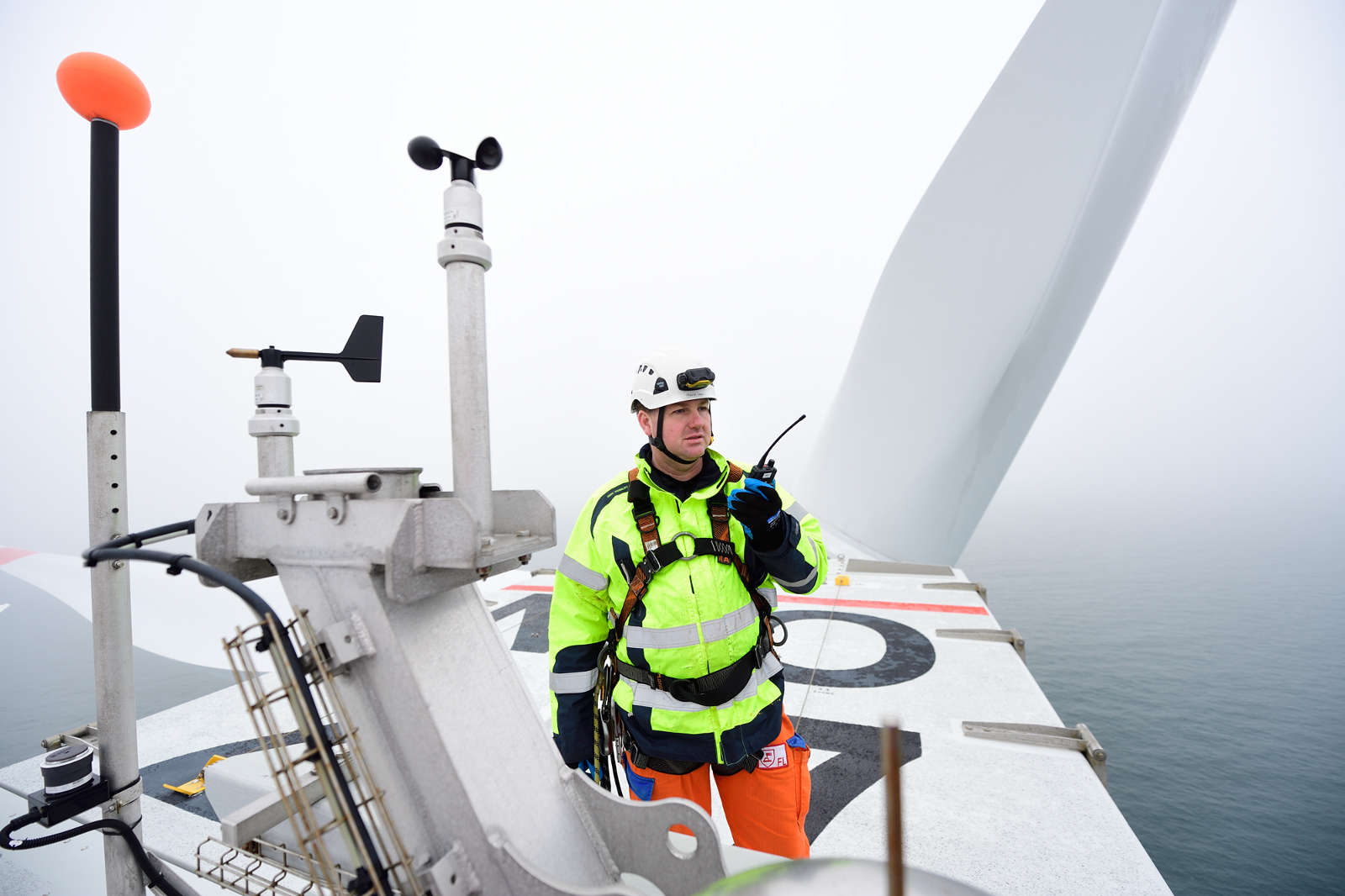 Ein Techniker in leuchtender Kleidung steht auf einer Windkraftanlage und spricht über ein Funkgerät in nebliger Umgebung.