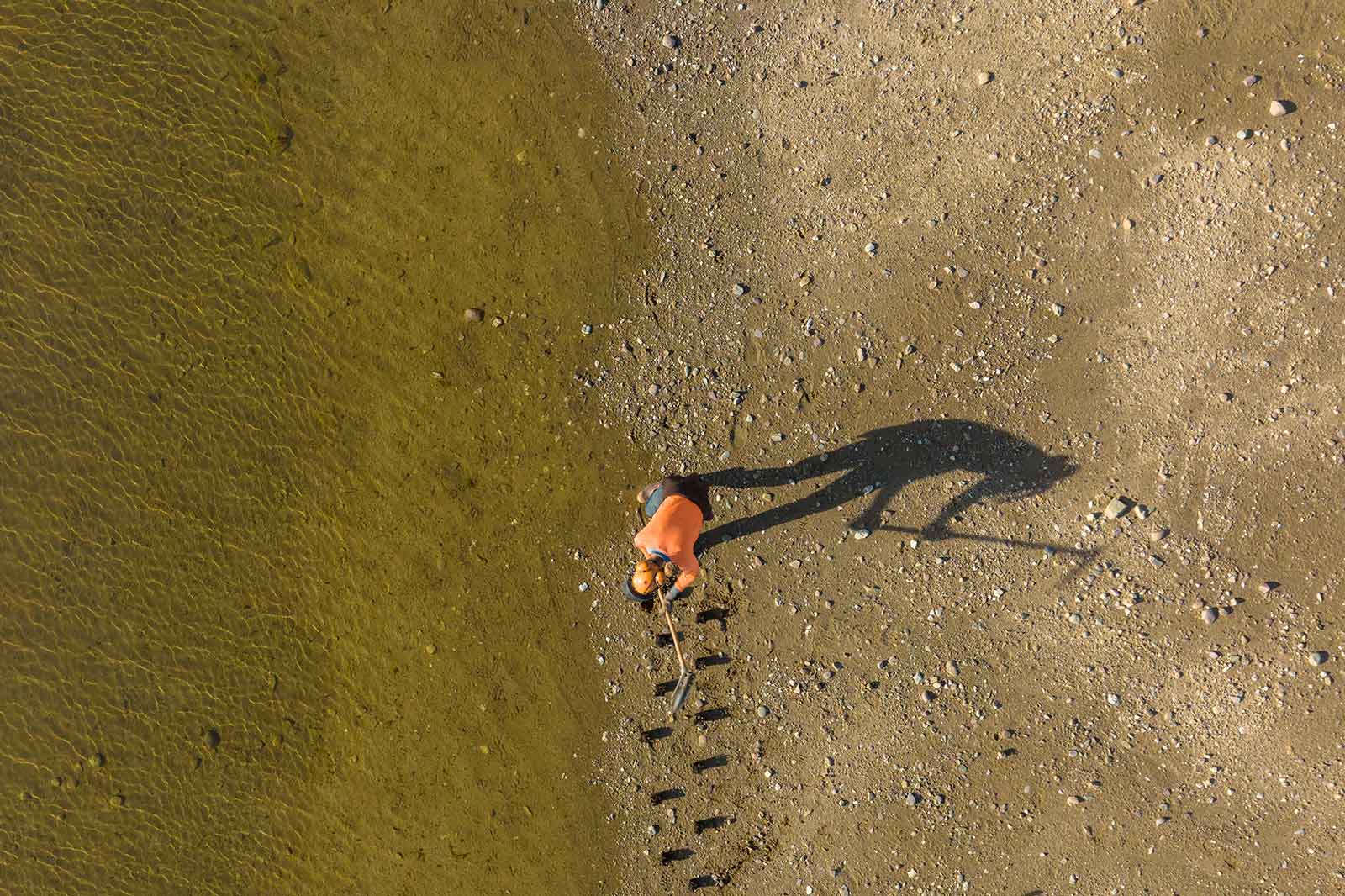 Eine Luftaufnahme einer Person in einem orangefarbenen Shirt, die in einem kiesigen Bereich nahe flachem Wasser gräbt.