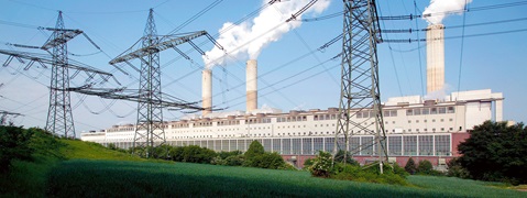 A power plant with smokestacks emitting steam, surrounded by greenery and high-voltage power lines.