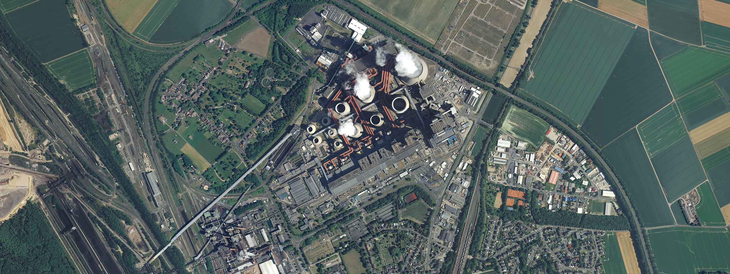 Aerial view of an industrial complex with water towers, smoking stacks, and surrounding fields and residential areas.