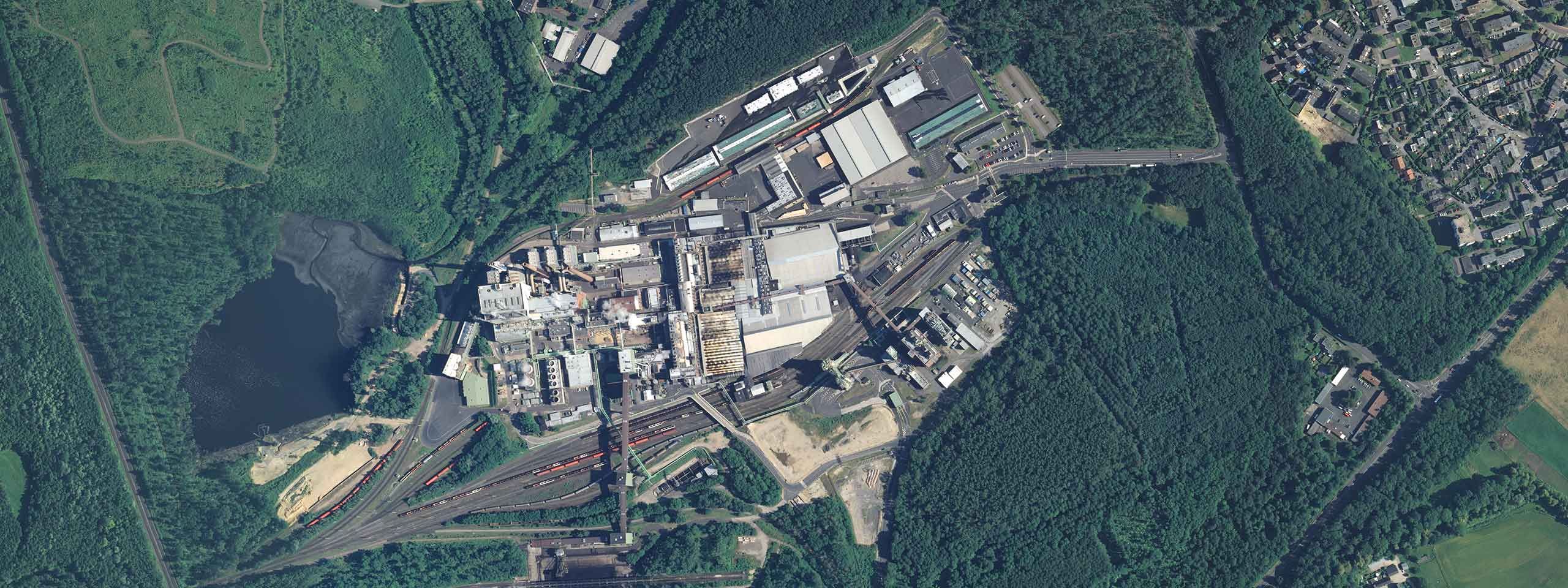 Luftaufnahme einer Industrieanlage, umgeben von Wald und einem kleinen Teich.