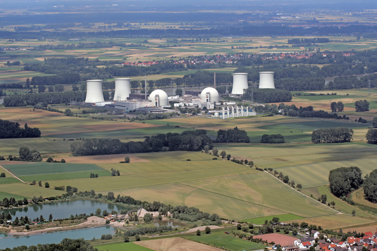 Aerial view of a nuclear power plant surrounded by fields and a small lake. Cooling towers and several buildings are visible.