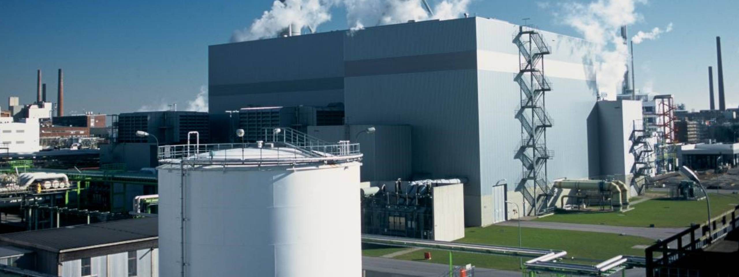 Eine Industrieanlage mit großen Gebäuden, Dampfwolken und hohen Schornsteinen im Hintergrund. Grüne Rohre sind sichtbar.